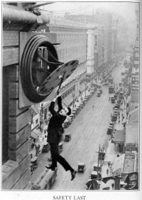  Harold Lloyd clutching the hands of a large clock as he dangles from the outside of a skyscraper above moving traffic